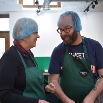 A person we support and staff member in the kitchen wearing aprons and hair nets preparing food and talking