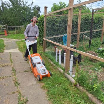 A person we support outside using a lawn mower to mow the grass wearing glasses and ear muffs