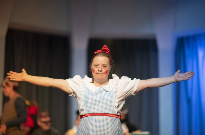 A person we support wearing a costume in a play, wearing a blue checkered pinafore and red bow