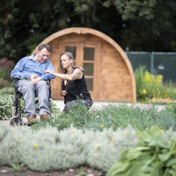 A person we support in a wheelchair and a staff member sitting in a garden together looking at a flower in Camphill Village Trust, UK charity