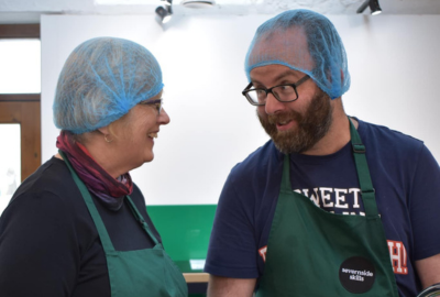 A person we support and staff member in the kitchen wearing aprons and hair nets preparing food and talking